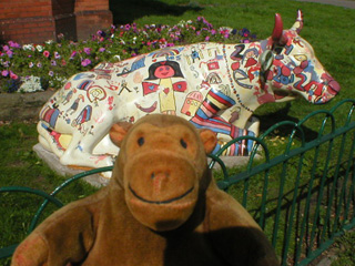 Mr Monkey in front of a reclining cow covered in bright pictures by children the guise of a shoe