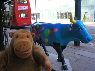 Mr Monkey with a cow decorated with balloons flying above a city skyline
