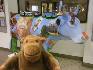 Mr Monkey in front of a cow decorated with scenes of Mancunian buildings and transport
