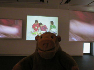 Mr Monkey looking at three video screens showing a family cooking