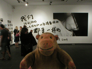 Mr Monkey looking across the gallery at a large photo with Chinese characters painted beside it