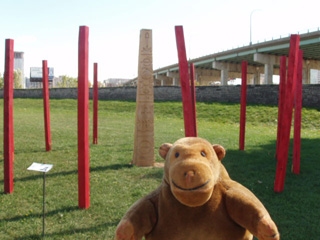 Mr Monkey in front of an installation with a carved shaft surrounded by red poles