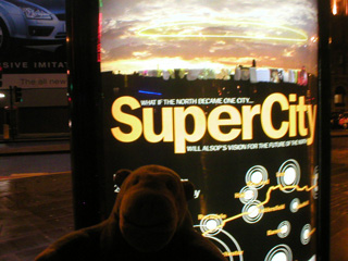 Mr Monkey beside a SuperCity advert in Piccadilly Gardens