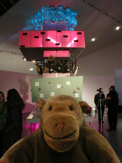 Mr Monkey in front of a high model with a green layer, a pink layer, and blue balloons